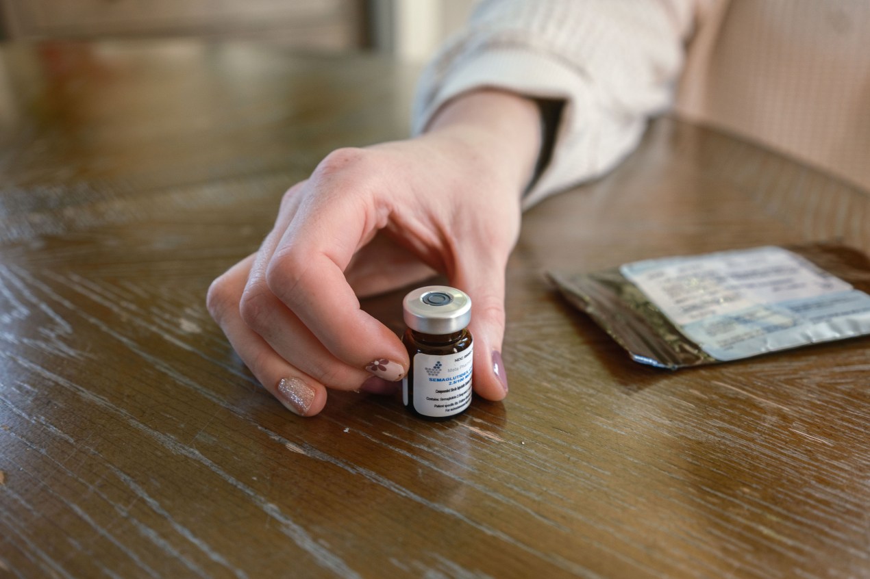 mcclain_04 A woman's hand holds a small vial of liquid GLP-1 medication on a table.