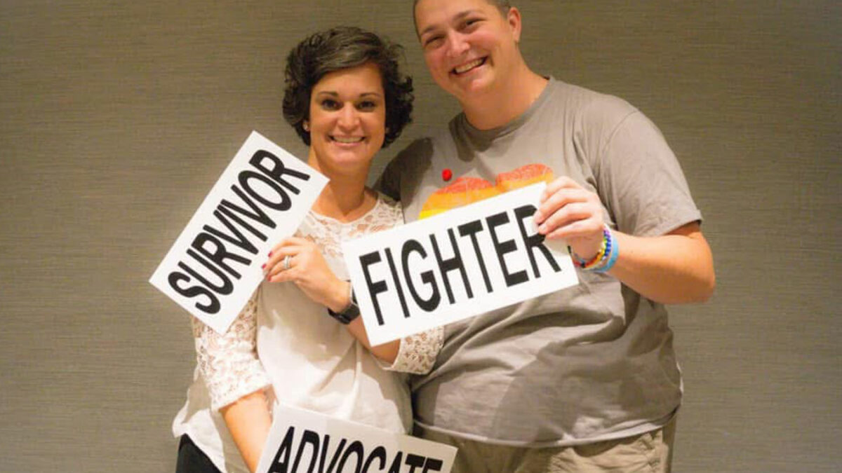 Two people pose for a photo together, smiling. The person on the left holds signs that read, "Survivor" and "Avocate." The person the right holds a sign that reads, "Fighter."