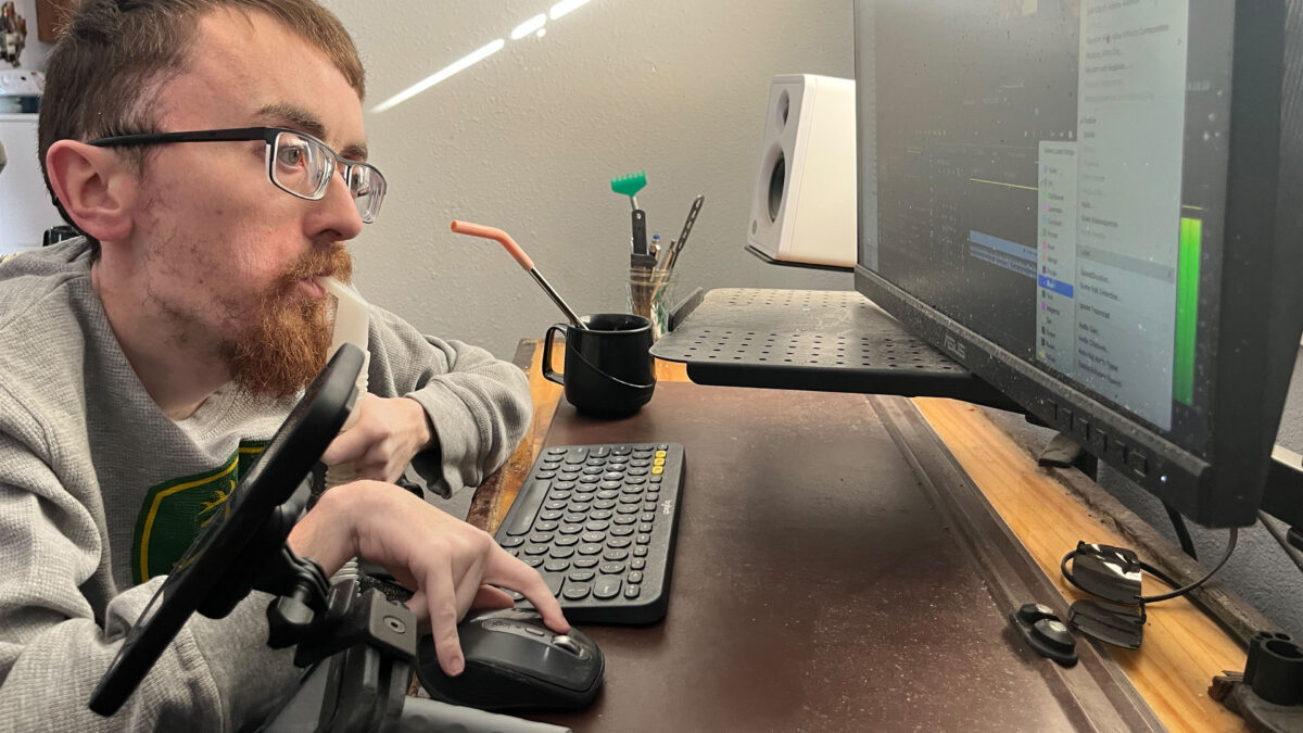 A photo of a man seated at a desk, working on a computer, breathing through a portable ventilator.