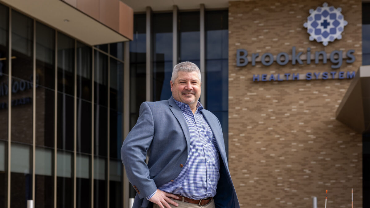 A man in business attire, with short-cut greying hair, stands in front of a hospital building. With his hands on his hips, he smiles. Behind him, building signage reads: "Brookings Health System"