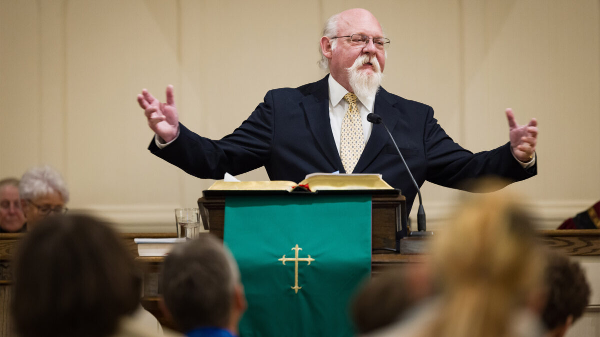 A reverend speaks at a lectern. People are seated in front of him, blurred in the foreground.
