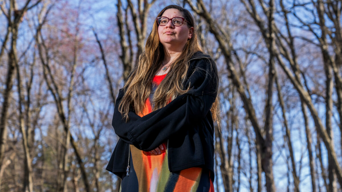 A woman stands outside in a wooded area.