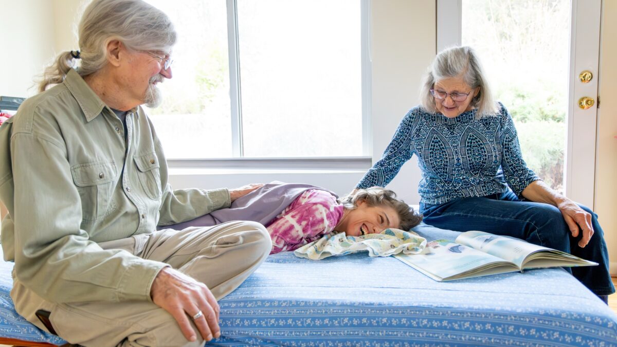 An older mother and father sit on a bed beside a smiling woman, laying down. The woman is living with a disability.