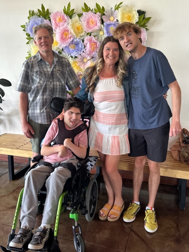 From left to right, four people smile for a photo as they stand in front of a flower mural: a man in a plaid shirt, a man in a pink shirt sitting in a wheelchair, a woman in a pink-and-white striped dress, and a man in a blue t-shirt