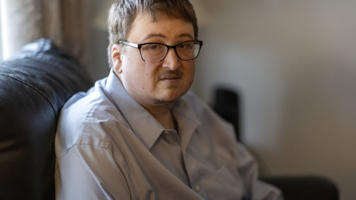 A man wearing glasses sits on a couch facing the camera.
