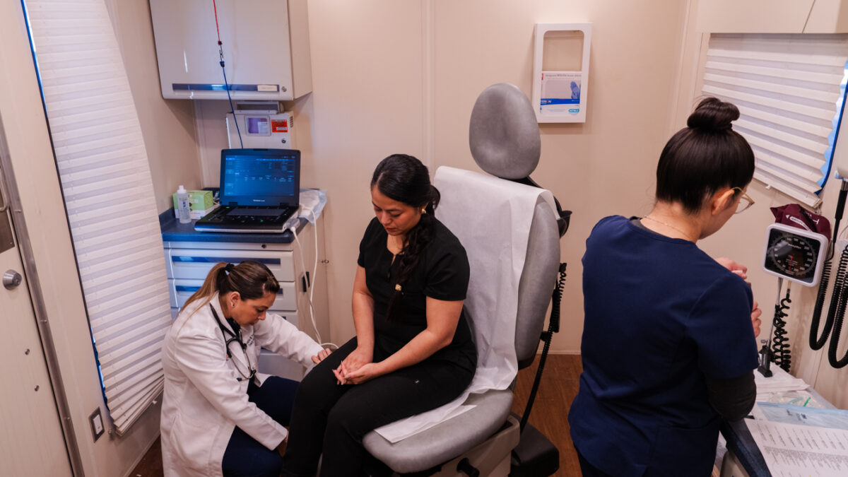 A photo of two medical professionals helping a women sitting in a chair inside a mobile clinic.