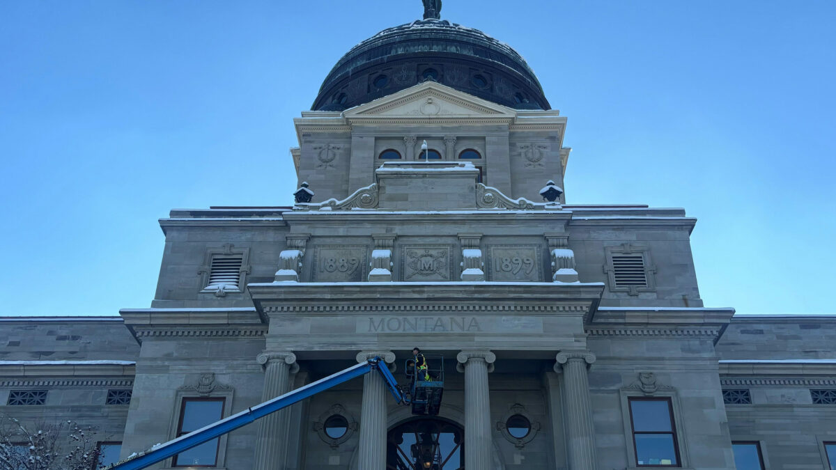 A photo of Montana's capitol with a few inches of snow. A construction worker in the foreground works on the building from a cherry-picker basket.
