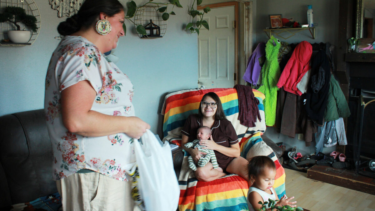 A doula stands next to a young mother sitting in a chair draped with colorful fabric. She holds a young baby in her arms.