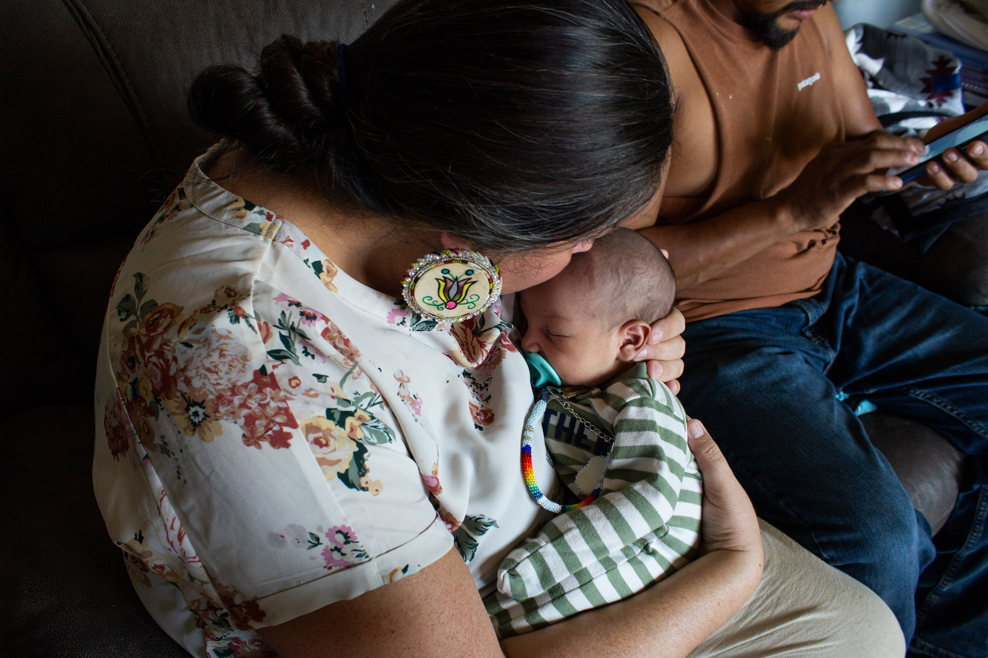 Misty Pipe is seen from the side. She kisses the forehead of a young baby. A man is seen behind her using his phone.