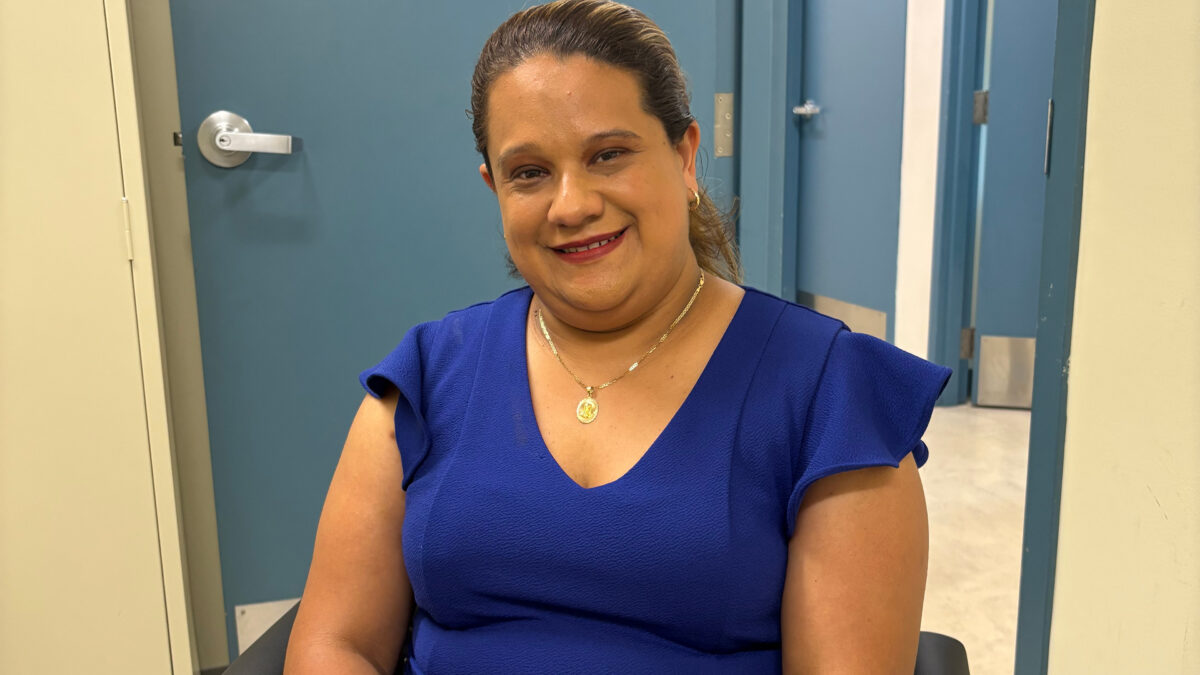 A woman in a blue tshirt with dark hair and red lipstick smiles at the camera. She is seated in front of a blue door