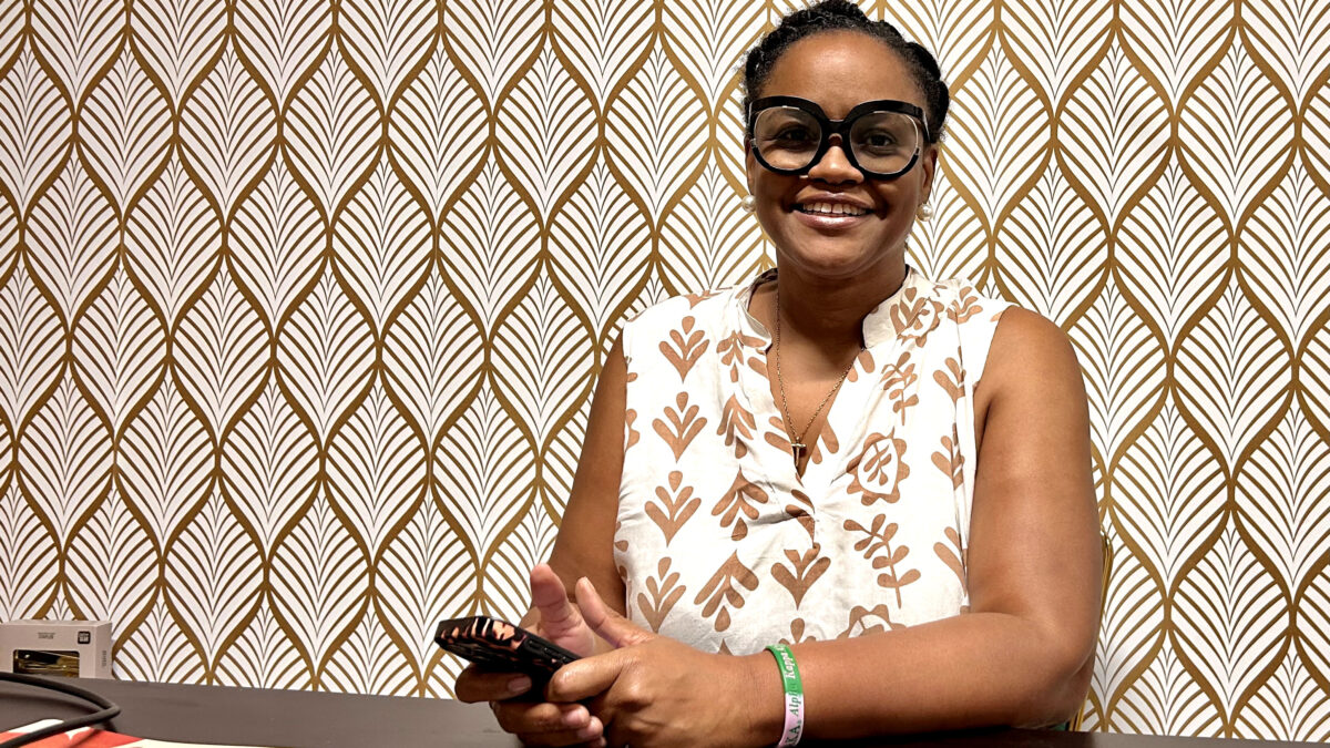 A woman wearing black-rimmed glasses and a white-and-brown patterned top sits at a table holding a cell phone while posing for a portrait