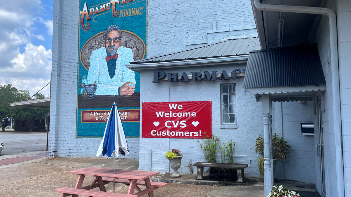 A photograph of the exterior of the Adams Family Pharmacy on a sunny day. There is a red sign out front that reads: "We Welcome CVS Customers!"