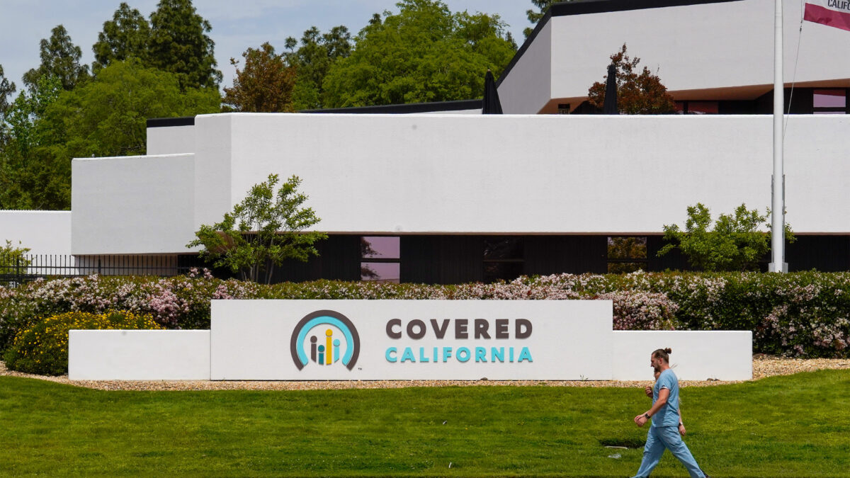 A sign in front of a building reads "Covered California." Two people walk along the sidewalk in front of the sign.