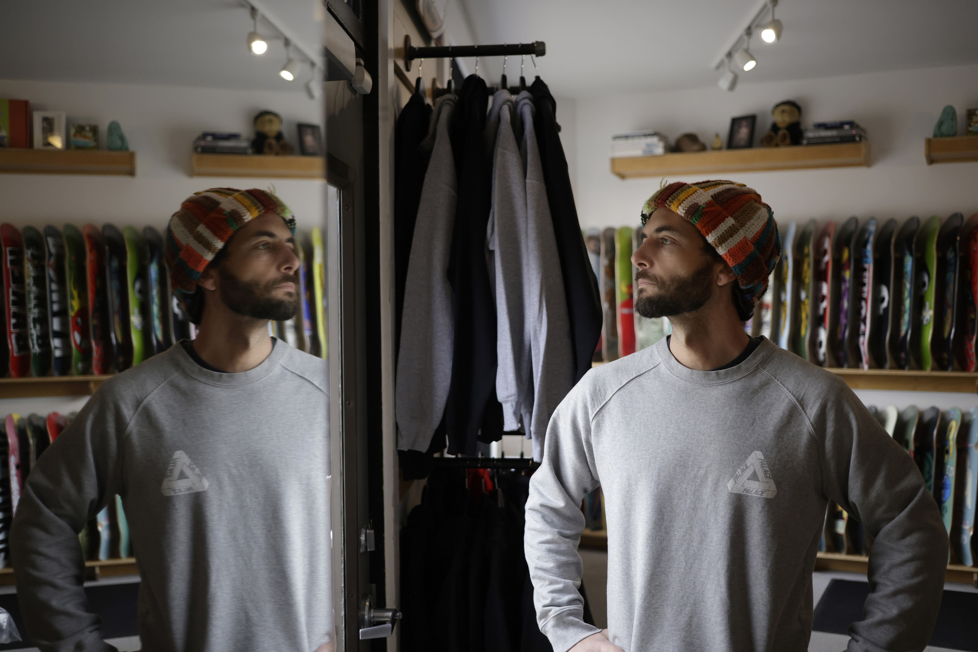 A man wearing a multicolor hat looks out the front door of a shop with skateboards on shelves behind him as the camera catches his reflection in the mirror