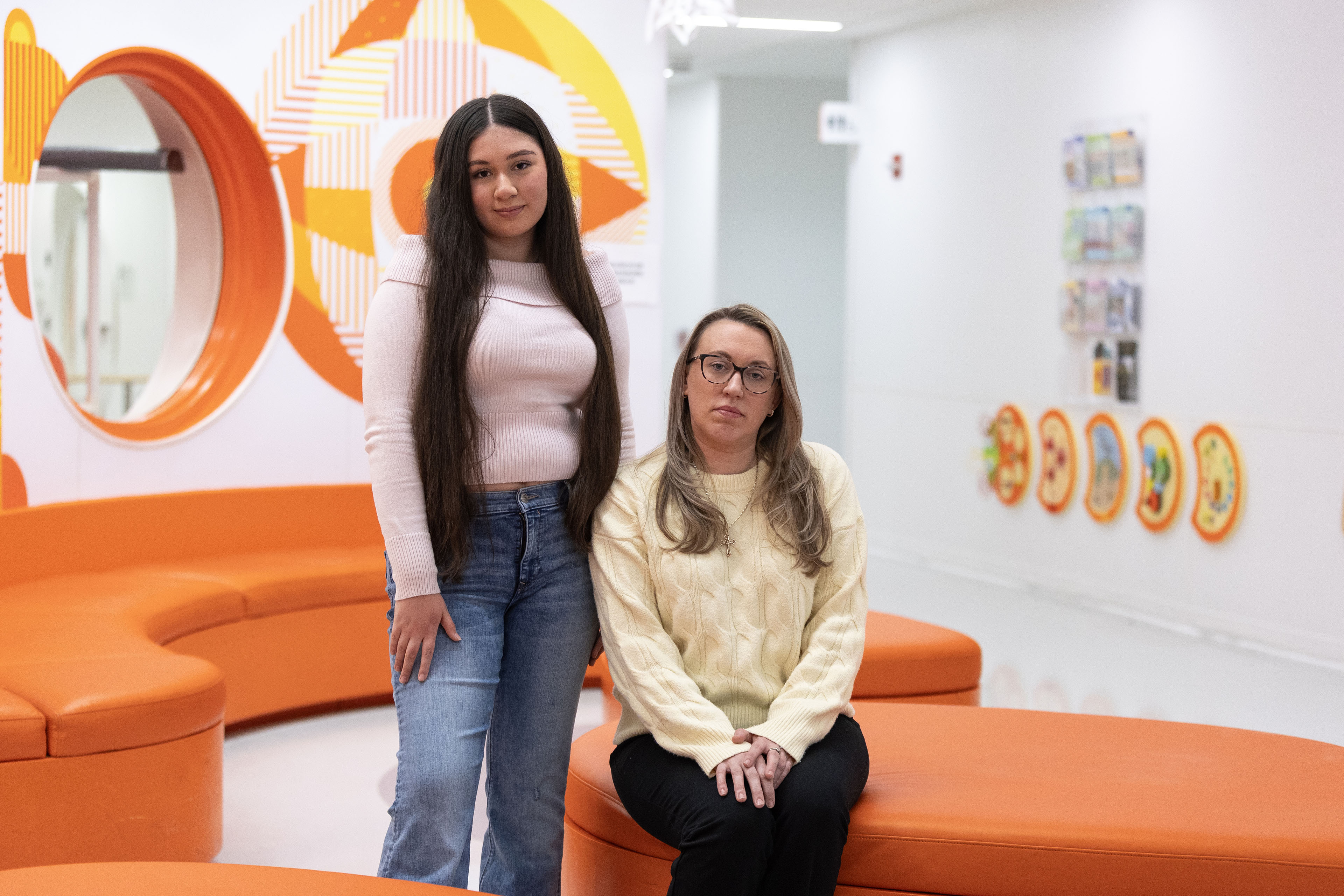 Two women pose for a portrait. On left, a woman with long brown hair, jeans and a sweater stands next to a woman with blond hair and glasses wearing a light yellow sweater