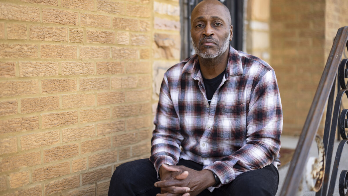 A man wearing a plaid button-up shirt sits on the front step of a home for a portrait