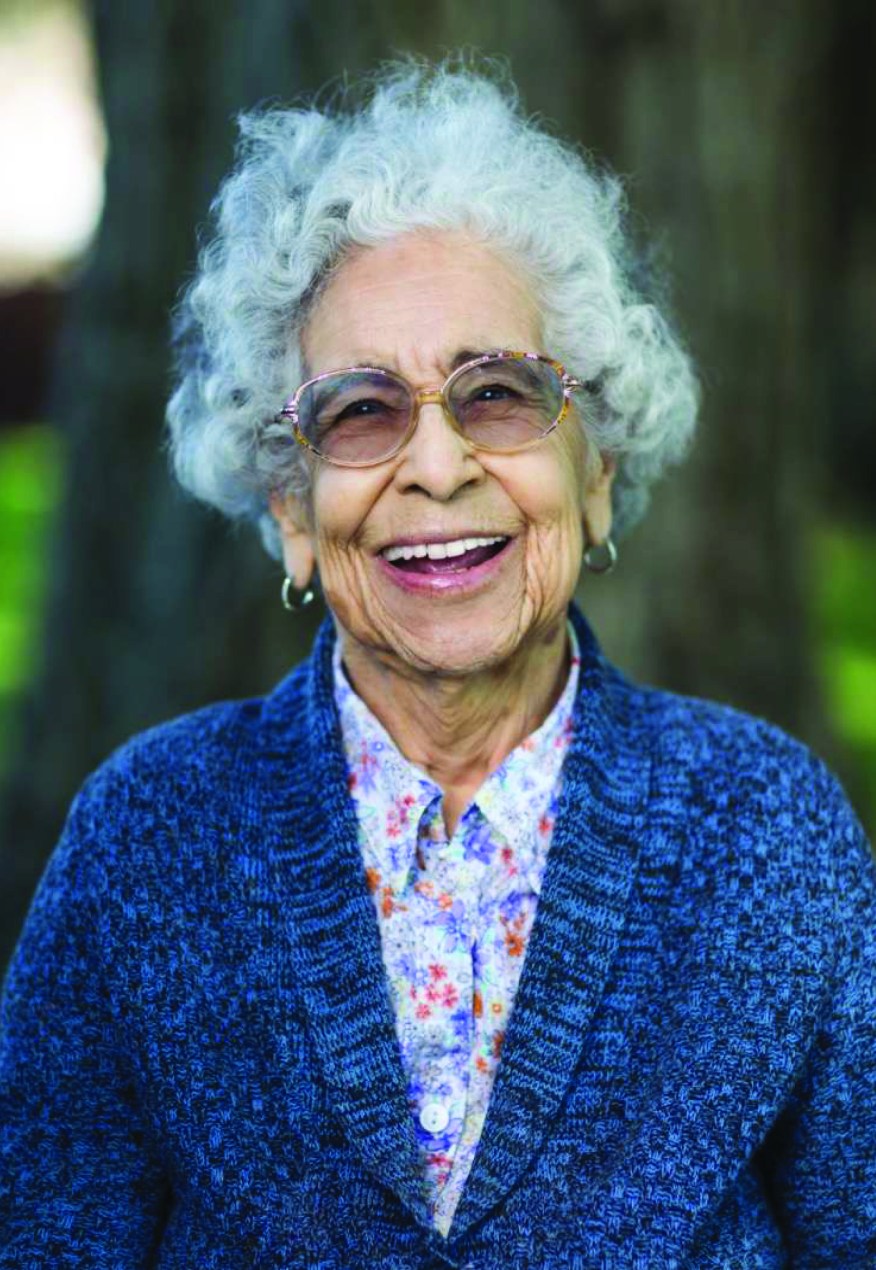 reit-mildred-hernandez-01-lo A woman with white hair and glasses, wearing a blue sweater and a floral shirt, smiles for a portrait