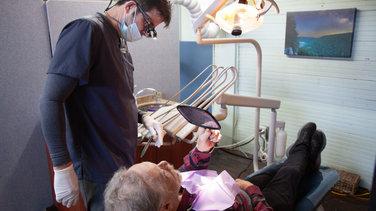 A photo of a dentist working with his patient, who is lying back in a dental chair, using a mirror to look at his teeth.