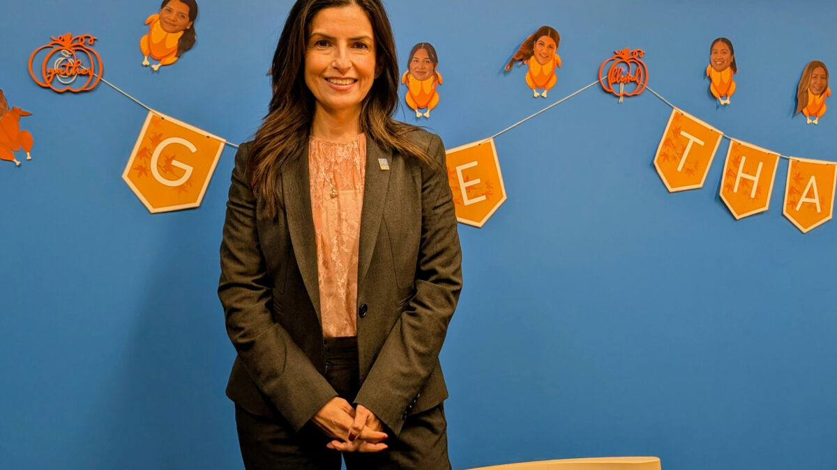 A photo of Martha Santana-Chin smiling in front of a wall painted blue. The wall appears decorated for Thanksgiving: a string of pennants spells out "Give Thanks." Images of employees' heads are superimposed on turkeys.