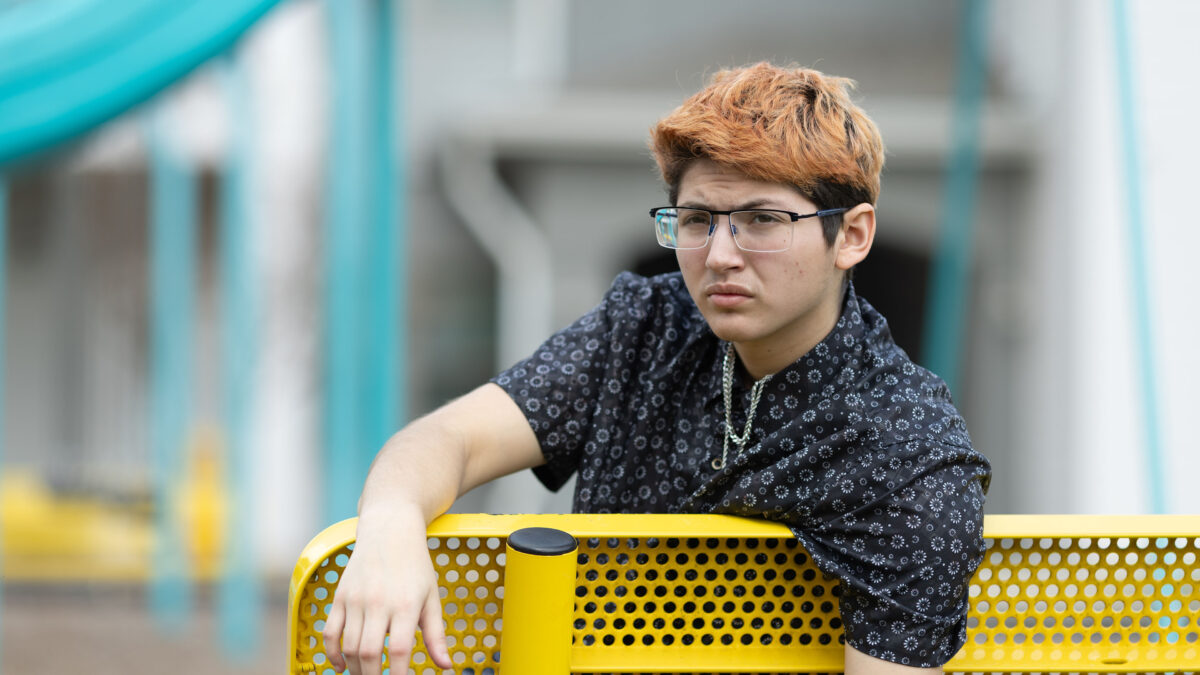 A portrait of a 20-year-old man wearing a short sleeve button up shirt and glasses leaning over the back of a yellow park bench.