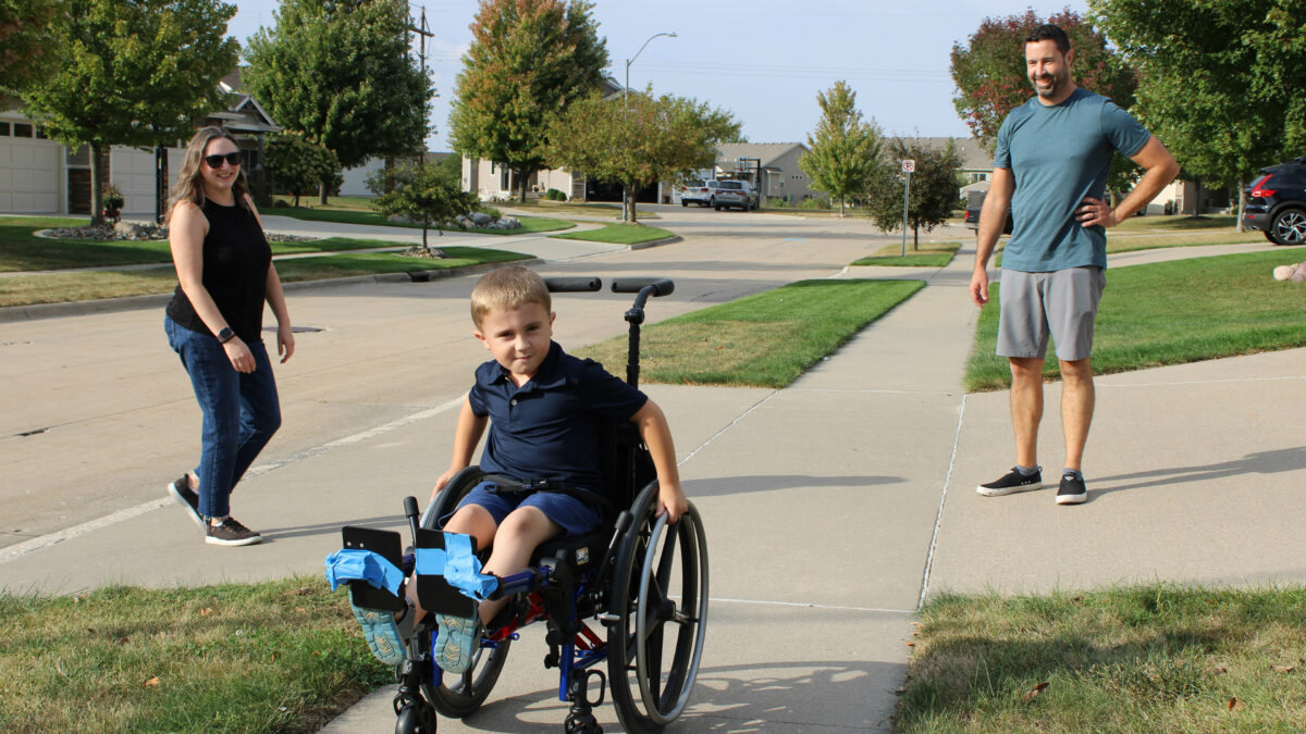 A young child in a wheelchair plays in a sidewalk with his mother to his left and his father to his right.