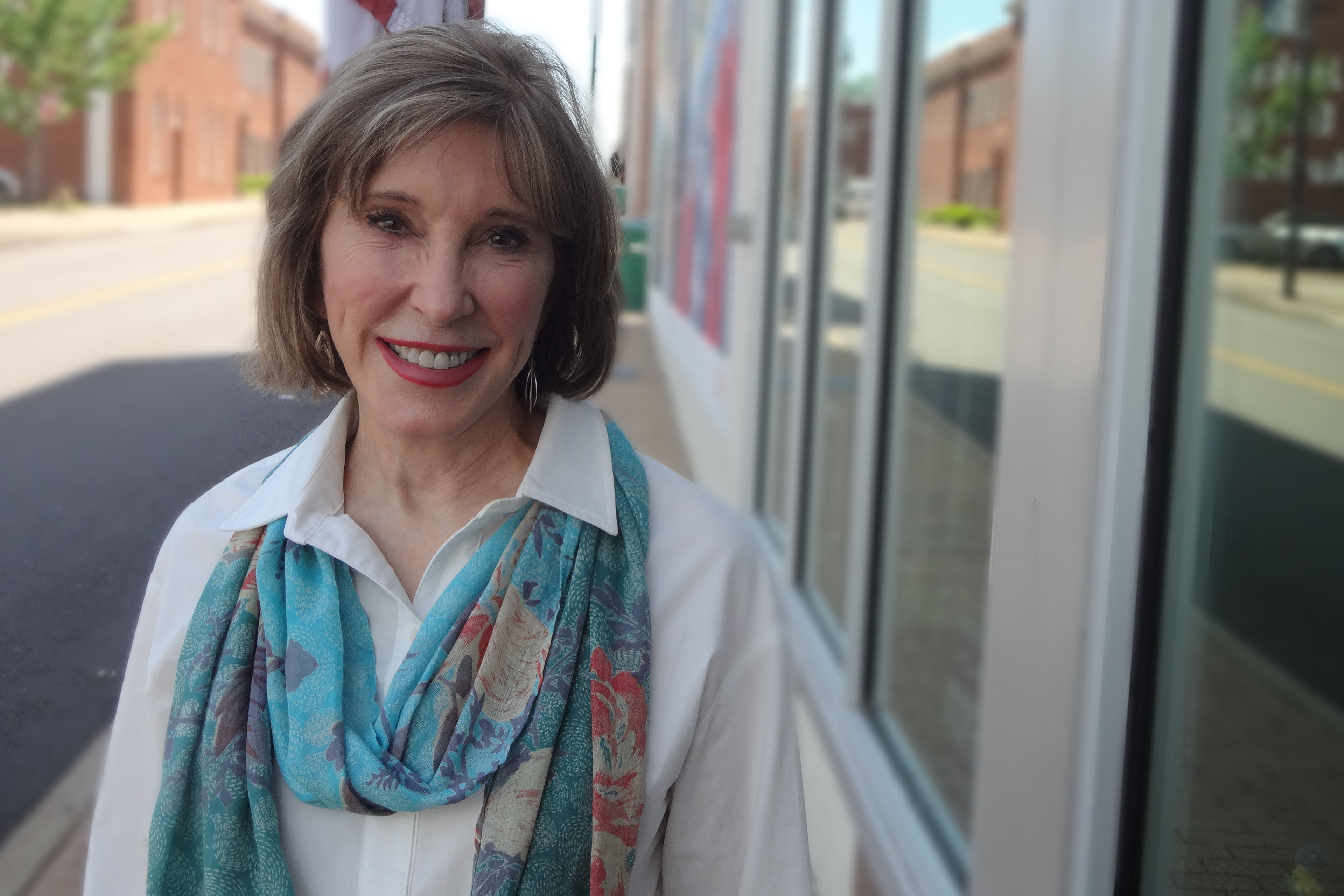Jill Holland stands outside a large building with glass windows. She wears a white blouse with a colorful scarf and smiles towards the camera.