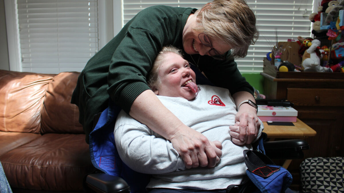 A mother leans over her child, who is in a wheelchair. They are both smiling.