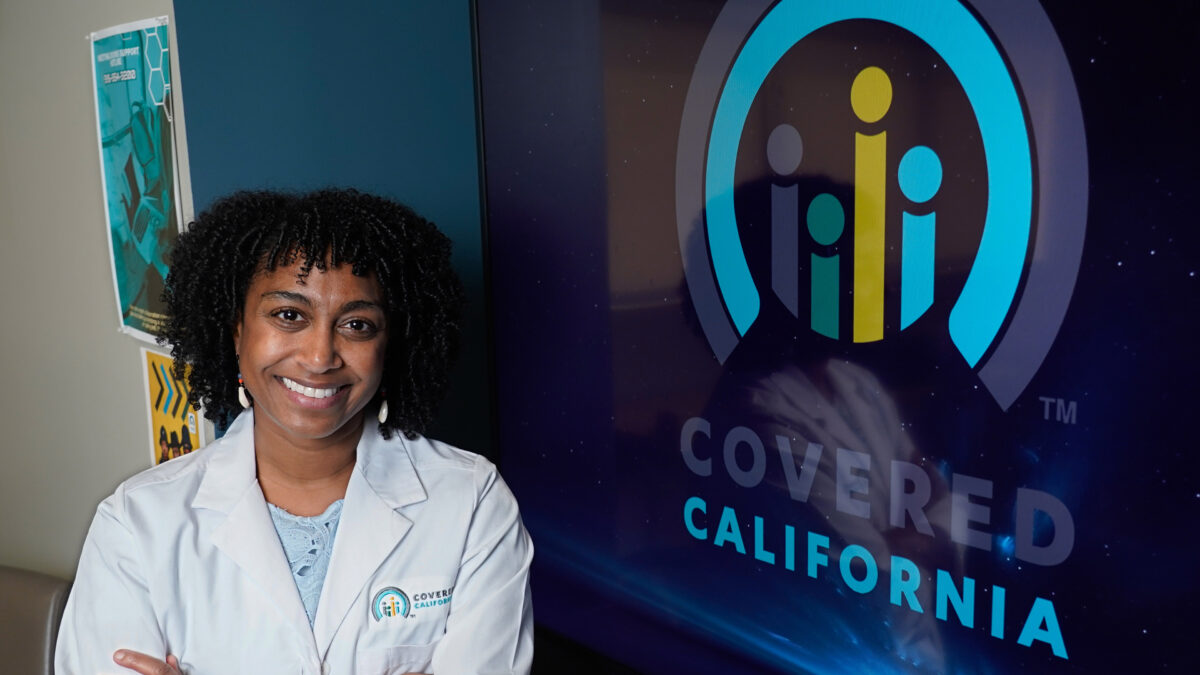 A photo of a woman in a white lab coat posing by a screen with the Covered California logo.