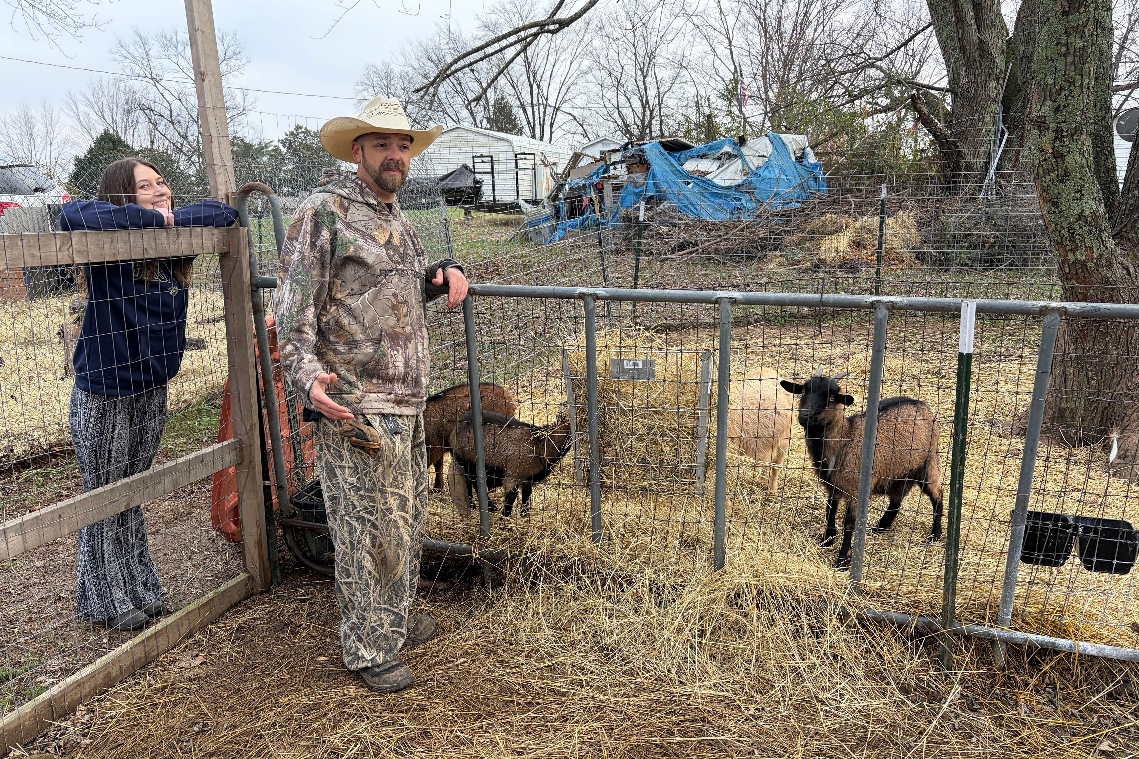 sory1_3840x2560web A man and a woman lean against the fences of a fenced-in area with straw on the ground and four visible goats. The woman with straight dark hair wears a dark blue sweatshirt with striped pants and smiles at the camera. The man with a beard wears a straw hat, camouflage sweatshirt, and camouflage pants is in the middle of talking and looks a something off-camera.