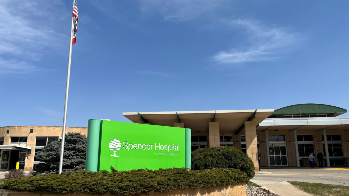 A photo of a hospital exterior with a neon green sign in front of it that reads, "Spencer Hospital, healthier together."
