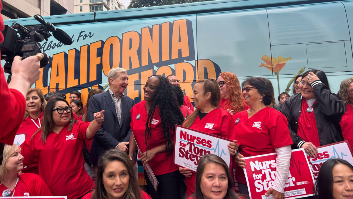 A group of people — most wearing red shirts — mingle in front of a bus with the words "All Aboard For A California You Can Afford" on its side.