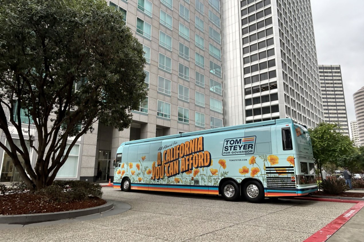 steyer_03_3840x2560web A bus with the message "All Aboard For A California You Can Afford" and "Tom Steyer for Governor" on its side is parked outside tall buildings.