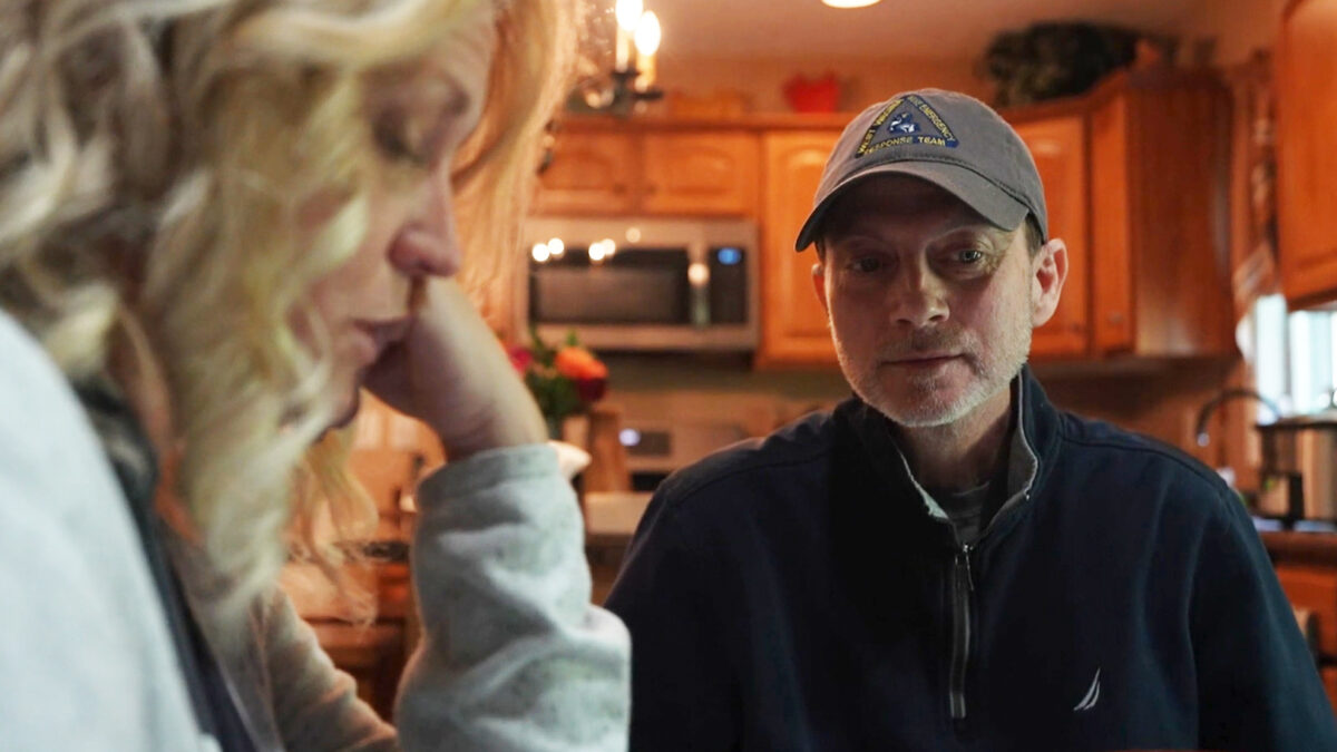 A husband sits at the kitchen table. His wife is seen close on the left of the frame. She is blurred slightly as the camera is focused on the husband.