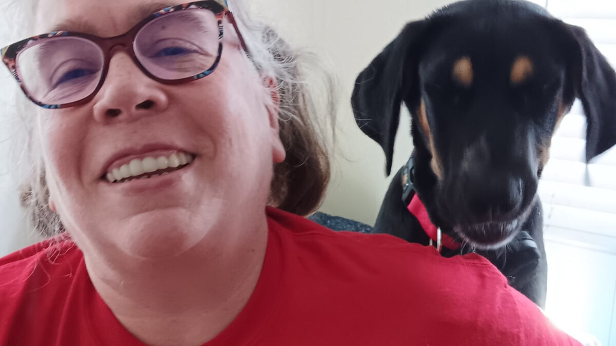 A woman in a red shirt and glasses smiles at the camera as a dog behind her looks over her left shoulder.