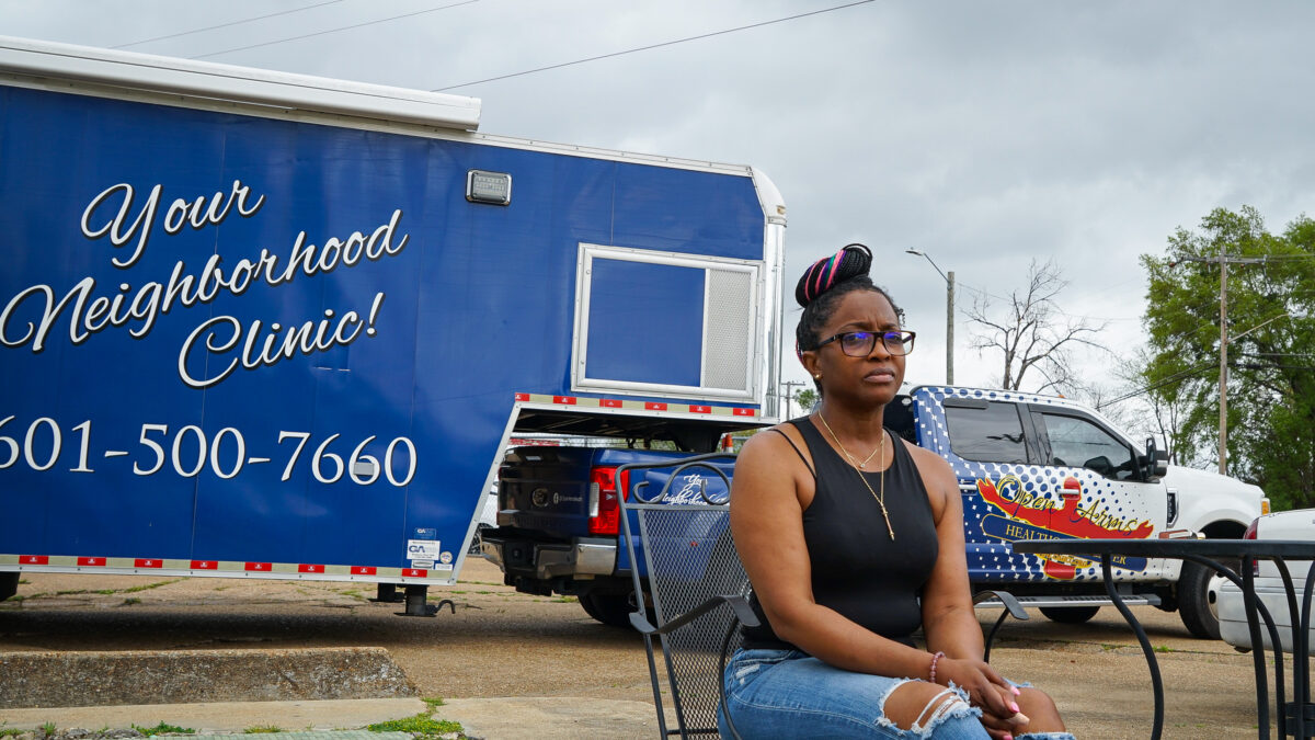 A woman sits in a folding chair in front of a blue van.
