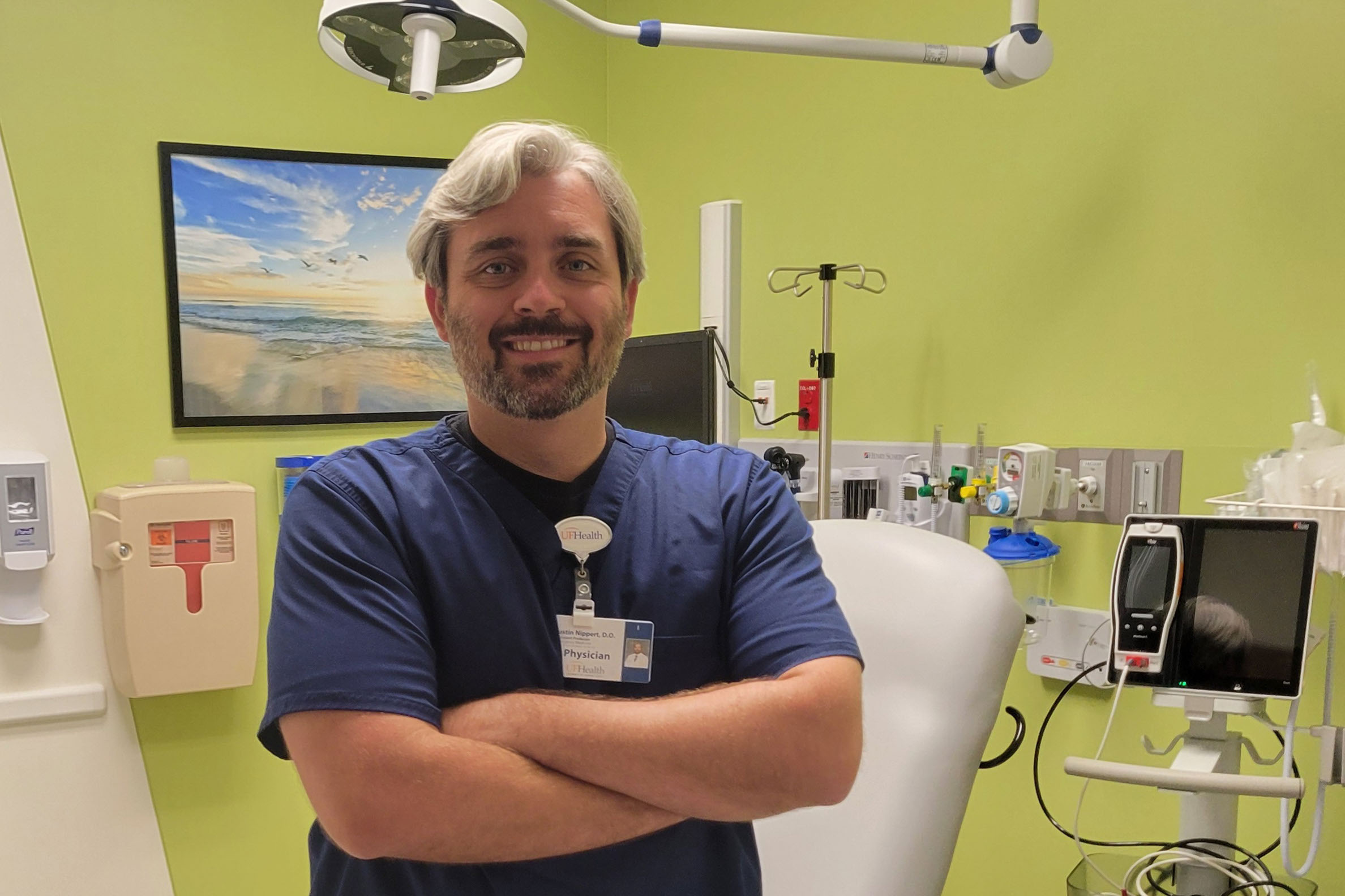 urgenters_06 Justin Nippert, an emergency physician, is standing in a medical room. He has his arms crossed and smiles broadly, facing the camera.