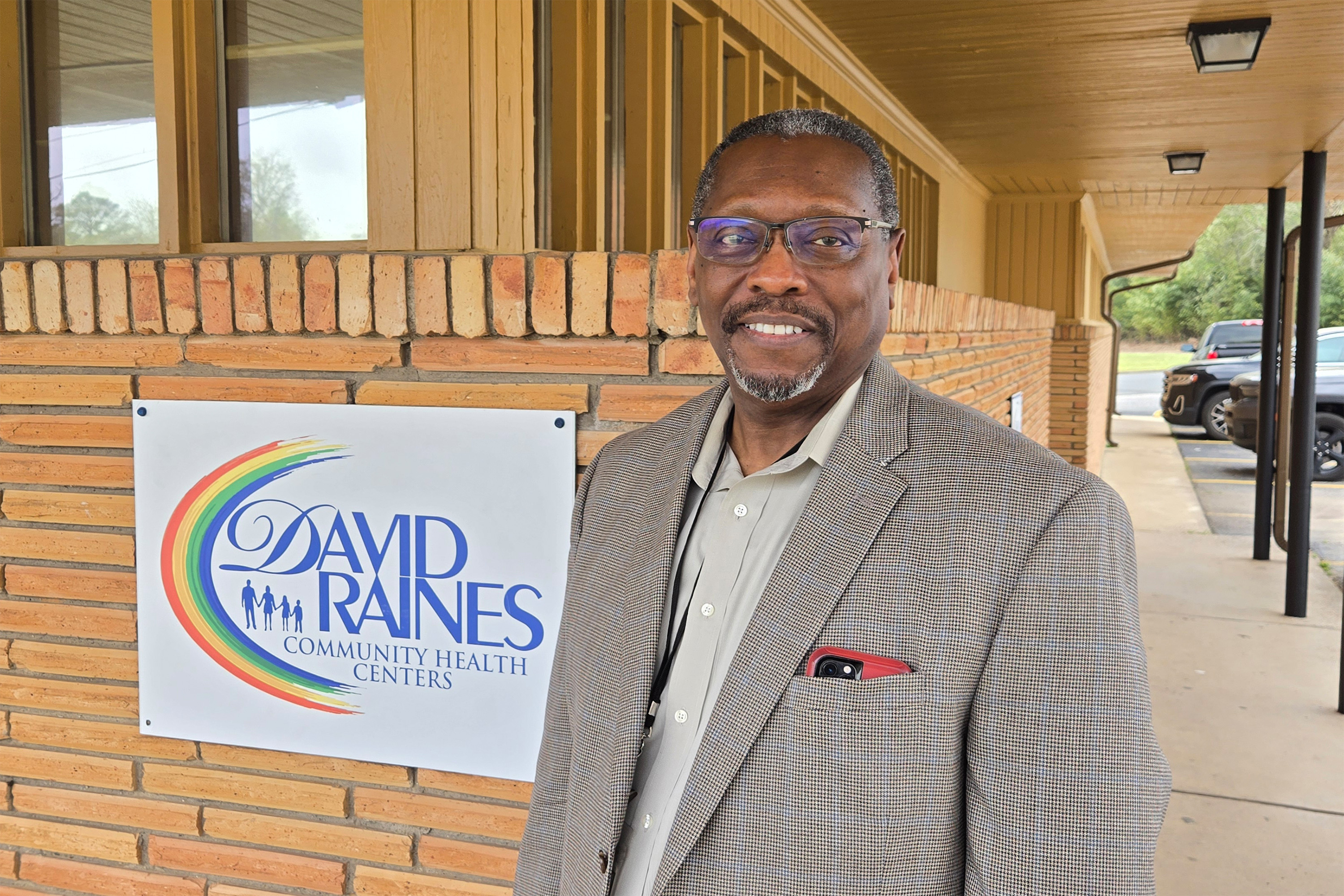white_david_raines A photo of a man in a suit smiling for a photo next to a sign that reads "David Raines Community Health Centers."