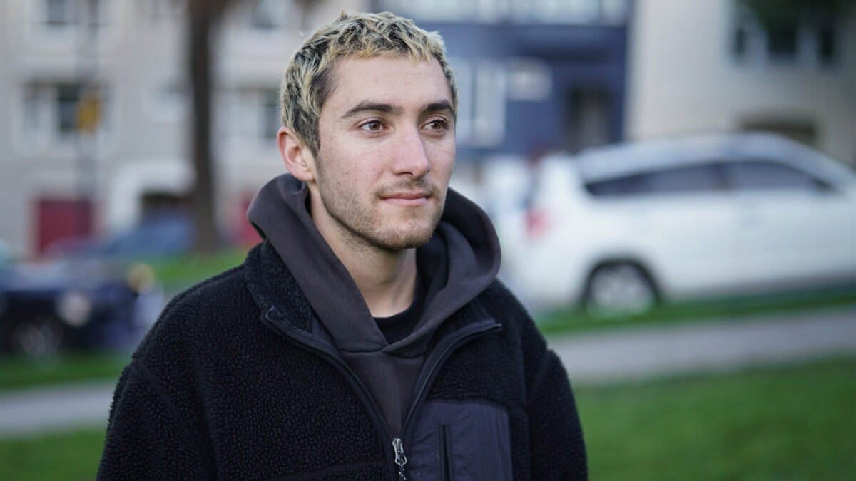 A photo of a young man posing for a portrait outside. A white car drives up the street behind him.