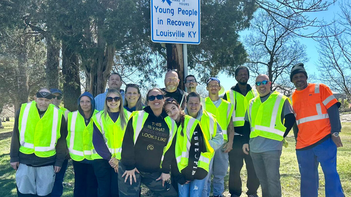 A photo of a group of people in reflective vests standing in front of a An Adopt a Highway sign that reads, "Young People in Recovery, Louisville, KY."